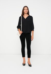 Black, loose-fitting blouse with a V-neck, paired with black, slim-fit pants. Model holds a small white handbag and wears black flats.
