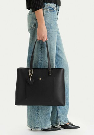 Persona con vaqueros azules y zapatos negros puntiagudos sosteniendo un gran bolso tote negro con un colgante en forma de corazón.