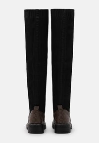 Botas negras de canalé hasta la rodilla con contrafuertes de cuero marrón con estampado y suelas negras gruesas, mostradas desde la parte trasera sobre un fondo blanco.