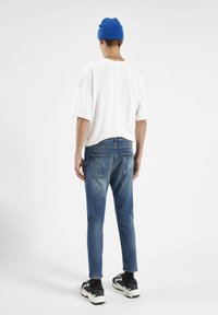 Camiseta blanca oversized, gorro azul, vaqueros ajustados azules con efecto deslavado, zapatillas negras con acentos rojos, collar de cadena plateado.