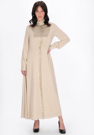Kobieta o ciemnych włosach, ubrana w długą beżową sukienkę na guziki i czarne szpilki, stojąca z jedną ręką na biodrze na białym tle.