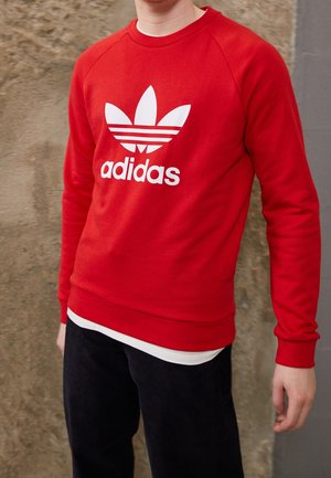 Osoba oblečená v červené mikině Adidas s bílým logem, černé kalhoty, stojící před texturovanou kamennou zdí.