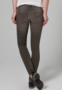 Jeans ajustados en marrón apagado, con una textura suave, costuras en contraste y dos bolsillos traseros con detalles de costura. Bailarinas visibles abajo.