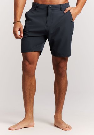 Man draagt zwarte korte broek tot halverwege de dij, staat blootsvoets, met één hand in de zak en de andere langs zijn zij rustend.