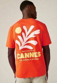 Kätevalt oranž puuvillane T-särk, millel on valge kunstiline splash-kava ja kollane kiri "Mediterranean CANNES THE FRENCH RIVIERA". Lühikesed varrukad.