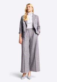 Gray and blue patterned blazer with large buttons, paired with wide-leg trousers. Underneath, a white ruffled blouse. Blue pointed heels.