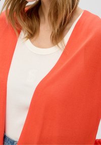 Korallfärgad kofta i mjukt material, med öppen front och lös passform, lagd över en vit linne med en broderad logotyp.