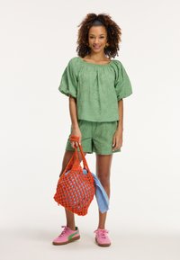Completo due pezzi verde con texture, maniche a palloncino e pantaloni corti, abbinato a una borsa intrecciata arancione brillante e sneakers rosa con accenti verdi.
