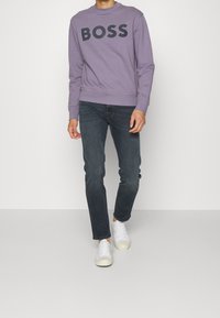 Felpa lilla con la scritta "BOSS" in grandi lettere nere, abbinata a jeans scuri e sneaker bianche. Presenta un collo rotondo e polsini a coste.