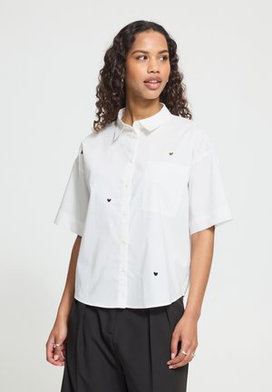 Jeune femme aux cheveux bouclés porte une chemise blanche à manches courtes boutonnée avec de petits cœurs noirs et marron et un pantalon noir.
