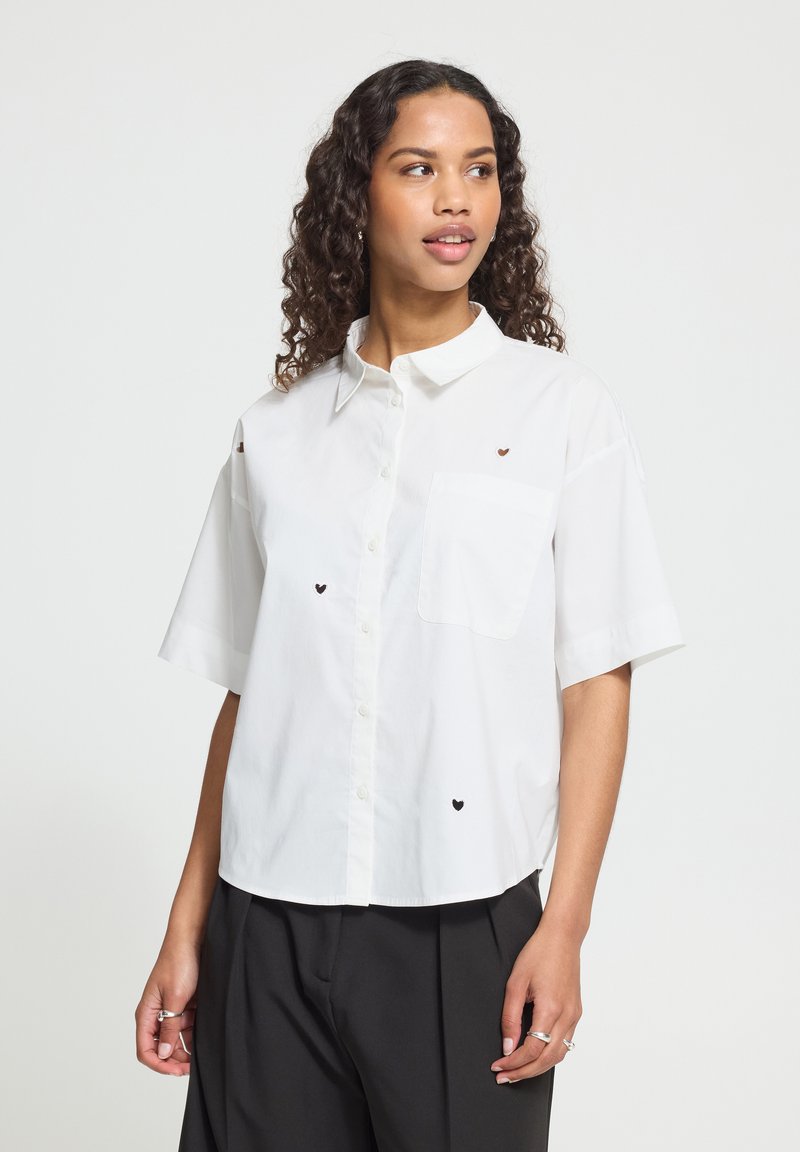 Jeune femme aux cheveux bouclés porte une chemise blanche à manches courtes boutonnée avec de petits cœurs noirs et marron et un pantalon noir.