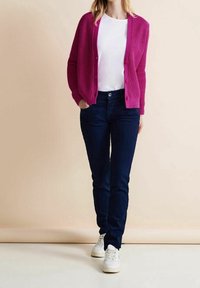 Ljusrosa ribbad kofta med knappstängning, bärs över en vit skjorta, kombinerat med mörkblå jeans och vita sneakers.