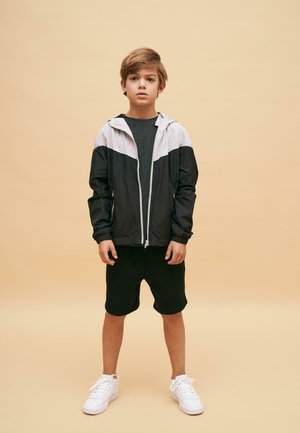 Giacca a zip nera e grigia chiara, pantaloni corti neri e sneakers bianche indossati da un bambino su uno sfondo neutro. Stile casual sportivo.