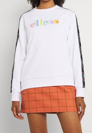 Osoba nosząca biały sweter z kolorowym logo "ellesse" i czarną taśmą z logo na rękawach, w połączeniu z pomarańczową mini spódniczką w kratkę.