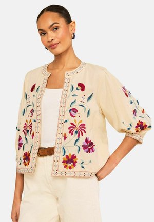 Naine kannab beeži lühikest jopet, millel on punane, roosa ja lilla lilleline tikand, valget toppi ja beežeid pükse pruuni vööga, käsi puusal.