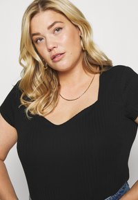 Zwart geribbeld topje met korte mouwen en een V-hals. Beschikt over een slim fit ontwerp en textuurstof. Gouden ketting als accent.