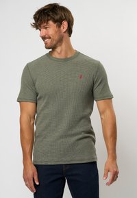 Tricou tricotat cu mânecă scurtă, de culoare verde, cu guler rotund și detaliu cu logo roșu în partea stângă a pieptului. Materialul pare texturat și respirabil.