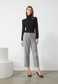 Pull en col roulé noir, ajusté ; pantalon à carreaux gris avec une coupe courte ; modèle portant des escarpins noirs à bout pointu ; arrière-plan minimaliste.