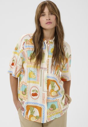 Frau mit langen braunen Haaren trägt ein buntes kurzärmeliges Hemd mit Obstmustern und steht mit den Händen in den Taschen einer beigen Hose.