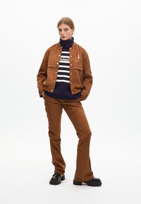 Hnědá semišová bunda se zacvakávacími knoflíky, v kombinaci s hnědými zvonovými kalhotami a námořnickým pruhovaným rolákem. Outfit doplňují černé robustní boty.