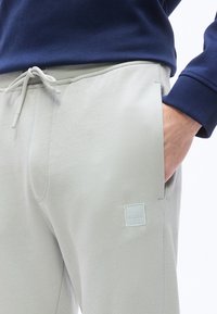 Άνδρας που φοράει ανοιχτό γκρι φόρμα με κορδόνι και λογότυπο BOSS, με το χέρι στην τσέπη, συνδυασμένη με μπλε ναυτικό μπλούζα μακρυμάνικη.