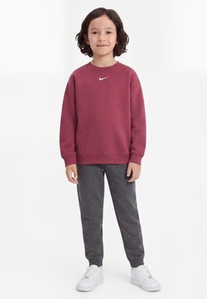 Dieťa s tmavými vlasmi po ramená, oblečené v bordovej mikine Nike, sivé tepláky a biele tenisky, stojace čelom vpred.
