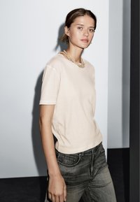 Kortærmet, lysebeige t-shirt lavet af blødt stof, parret med grå jeans. Den har en rund halsudskæring og en afslappet pasform uden synlige logoer.