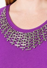 Lila Hemd mit einem dekorativen metallischen Kragen, der aus strukturierten Röhren und Perlen besteht und ein geometrisches Muster um den Halsausschnitt bildet.