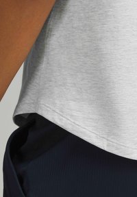 Hellgraues Sportoberteil mit abgerundetem Saum, kurzen Ärmeln und glatter Textur, über dunkellblauen, figurbetonten Hosen getragen.