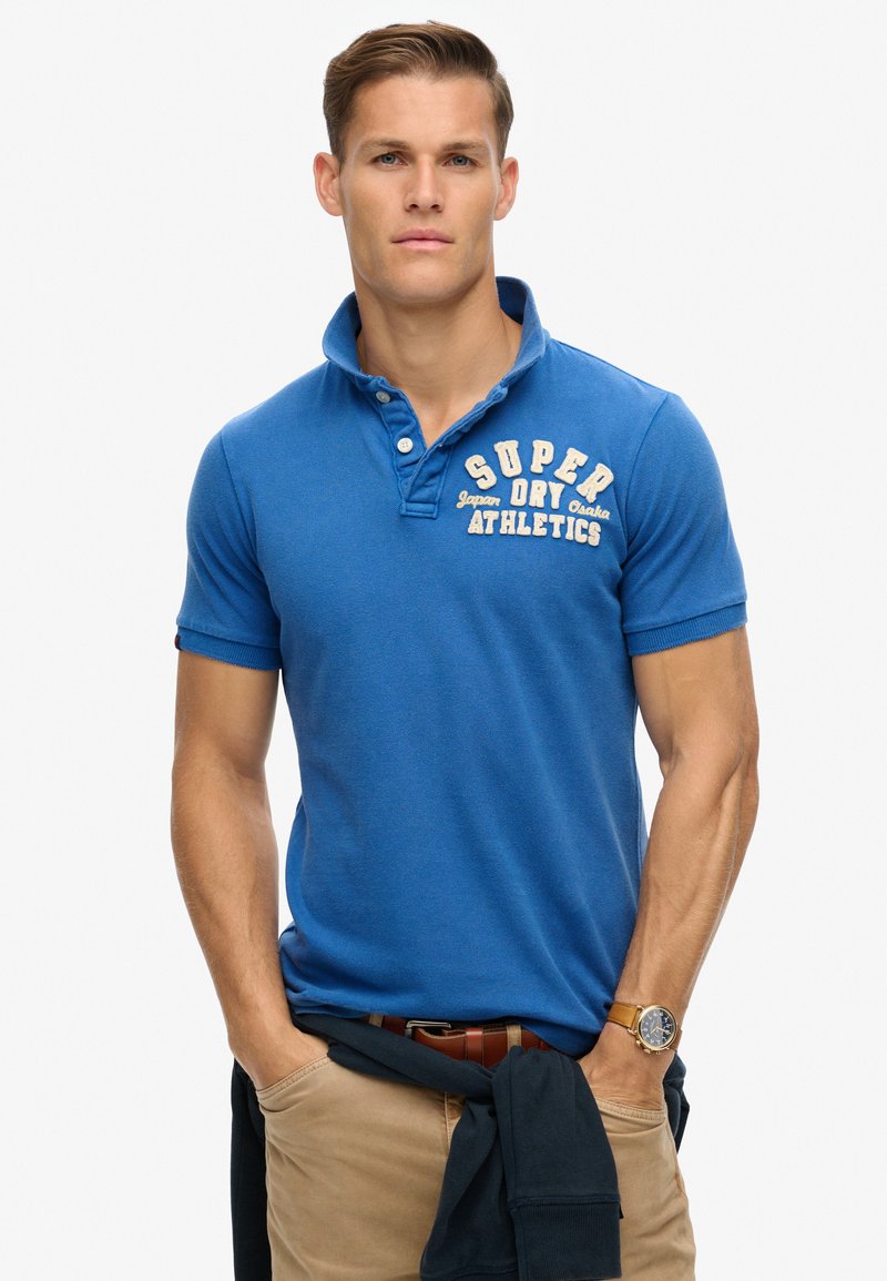 Blå polo t-shirt med korte ærmer, knaplukning og logo på brystet; iført med khaki bukser og en mørk trøje bundet omkring taljen.