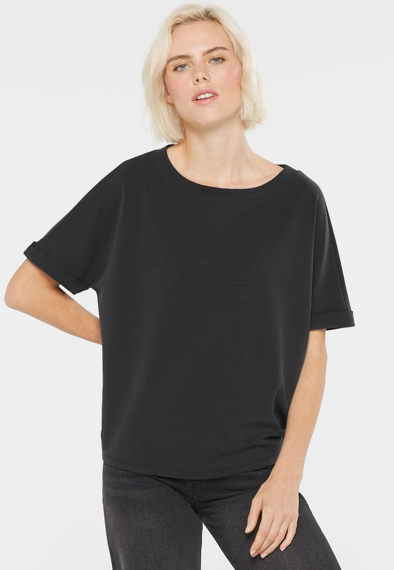 Blonde woman wearing a loose-fitting black short-sleeve shirt and dark jeans, standing with one hand on hip against a white background.