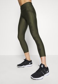 Olive green athletic leggings with a high waistband, featuring a black "WARRIOR" print along the side, paired with black sports shoes.