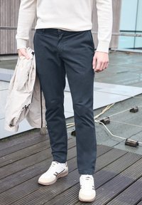 Tmavě modré chinos kalhoty s úzkým střihem, z bavlněného materiálu, s jemnou texturou, v kombinaci se světle béžovou bundou a bílými teniskami na dřevěné terase.