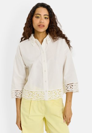 Femme aux longs cheveux bouclés portant une chemise blanche boutonnée avec des détails en dentelle et un pantalon jaune pâle, se tenant devant un fond blanc.