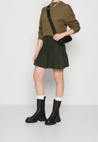 Botas negras de cuero hasta el tobillo con suelas gruesas, combinadas con una falda acampanada verde oscura y un suéter texturizado color oliva. Se lleva un pequeño bolso negro cruzado.