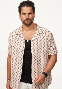 Kurzärmliges Hemd in Creme mit einem Zickzackmuster in Burgunderrot. Schwarzes Top darunter. Die Accessoires bestehen aus einer silbernen Halskette und einem Ring.