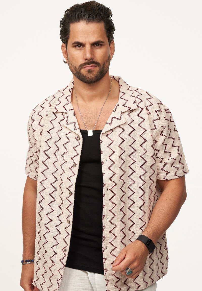 Kurzärmliges Hemd in Creme mit einem Zickzackmuster in Burgunderrot. Schwarzes Top darunter. Die Accessoires bestehen aus einer silbernen Halskette und einem Ring.