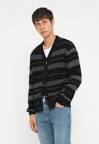 Cárdigan a rayas negro y gris con escote en V, botones al frente y puños acanalados. Usado sobre una camiseta blanca y combinado con jeans de mezclilla azul claro.