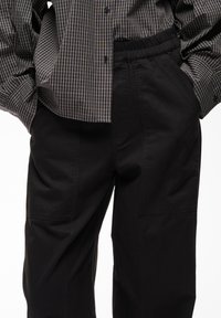 Pantalon noir à coupe ample avec grandes poches frontales et taille élastique, porté avec une chemise à manches longues à carreaux rentrée.