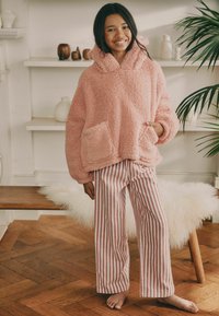 Gemütlicher rosa Fleece-Hoodie mit einer Kängurutasche und einer großen Kapuze, kombiniert mit weit geschnittenen gestreiften Hosen in sanften Pastellfarben.