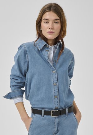 Mladá žena s hnědými vlasy, oblečená v modré džínové košili, pruhovaném tričku, černém pásku a modrých džínách, s rukama v kapse, neutrální výraz.