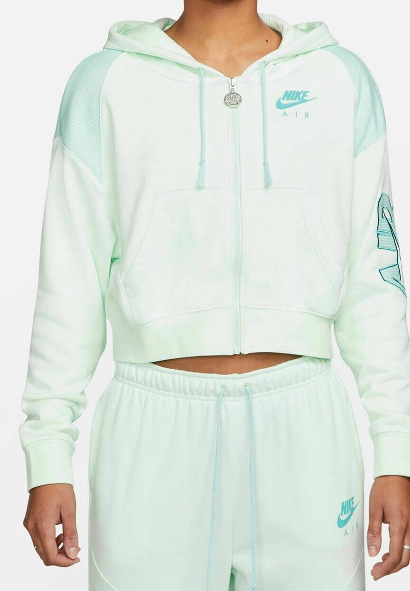 Kobieta ubrana w miętowo-zieloną, krótko przyciętą bluzę Nike Air z zamkiem i dopasowane spodnie dresowe, ręce swobodnie opuszczone wzdłuż ciała, bez widocznego tła.