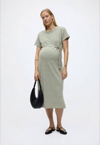 Έγκυος γυναίκα που φοράει φόρεμα με πράσινες και άσπρες ρίγες, κρατώντας μια μαύρη τσάντα, στέκεται μπροστά σε ένα απλό φωτεινό φόντο.