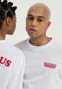 Deux personnes portant des chemises blanches avec le texte rouge "Delicious Refreshing Fruit" ; l'une fait face à l'avant montrant un tatouage sur le cou, l'autre est tournée de côté.