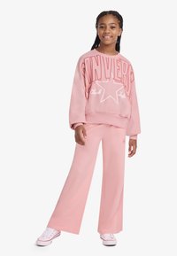 Ροζ φούτερ με μεγάλο γράμμα «CONVERSE» και σχέδιο αστέρι, σε συνδυασμό με ροζ παντελόνια σε χαλαρή γραμμή, στυλιζαρισμένα με λευκά αθλητικά παπούτσια.