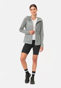 Giacca impermeabile grigio chiaro con cappuccio, tasche con zip e dettagli riflettenti, indossata sopra una camicia bianca e pantaloni corti neri.