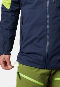 Giacca impermeabile blu navy con spalle a contrasto verde brillante; presenta tasche con zip e orlo regolabile; finitura liscia, tessuto leggero.