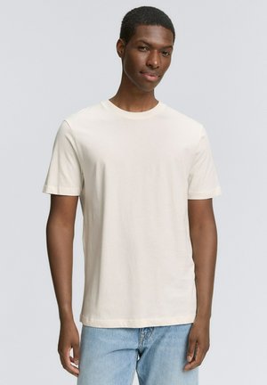 Cream-colored cotton t-shirt with short sleeves, a round neckline, and a relaxed fit. Worn with light denim jeans.