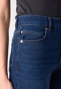 Tmavě modré džíny z denimu s mírně zvýšeným pasem, stříbrným kováním, designem s pěti kapsami a kontrastním šitím. Hladký povrch.