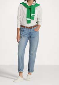 Witte button-up shirt, groene kabelgebreide trui om de hals, lichtblauwe denim jeans, bruine riem, witte sneakers. Casual outfit.